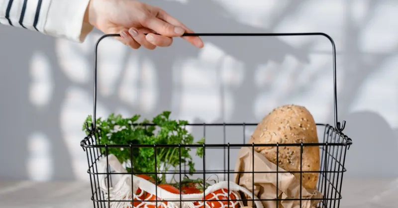 A hand holding a metal basket filled with eco-friendly groceries like bread, vegetables, and parsley.