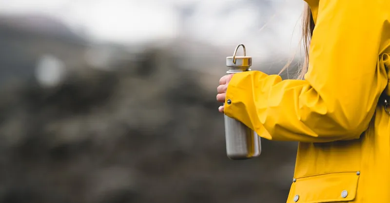 A person in a yellow raincoat holds a metal water bottle, set against a blurred Icelandic landscape.