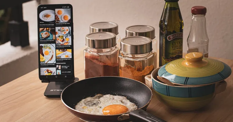Fried egg cooking with smartphone recipe guidance, spices and ingredients on a kitchen counter.