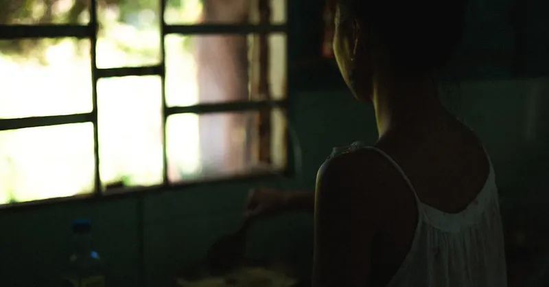 A woman stands by a window, cooking inside a dimly lit room, creating a serene mood.