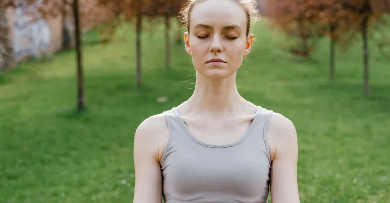 Young woman meditating outdoors in a lush green park during daytime, promoting mindfulness and relaxation.