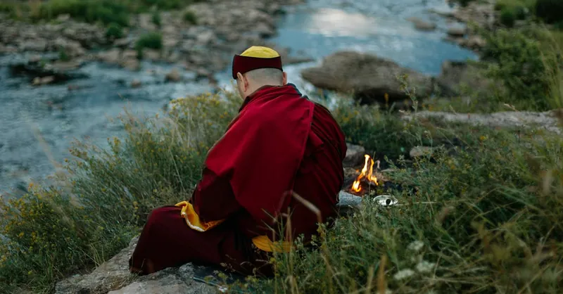 A serene scene of a Buddhist monk meditating by a river with a small campfire nearby.