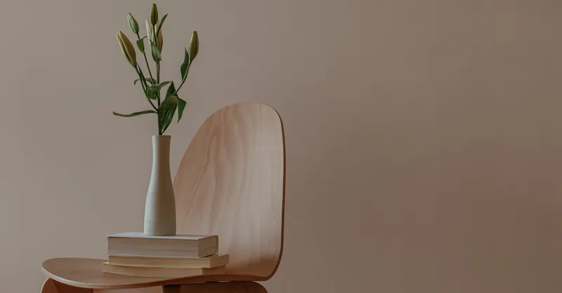 A minimalist setup with a wooden chair, vase, and stack of books indoors.