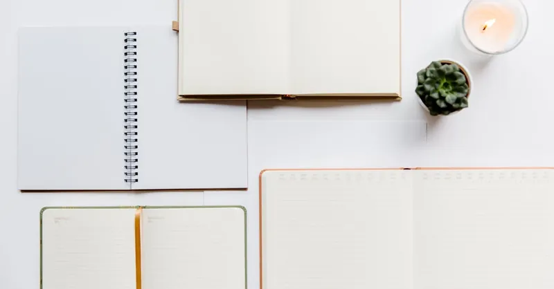A top view of a minimalist workspace showing open notebooks, a candle, and a small plant on a white background.