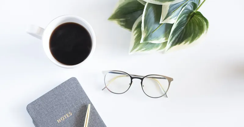 A minimalist workspace featuring a coffee cup, notebook, and eyeglasses on a white desk.