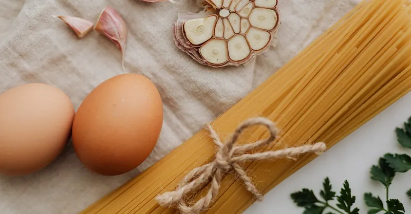 Flat lay of raw spaghetti, garlic, and eggs on linen, showcasing fresh ingredients.