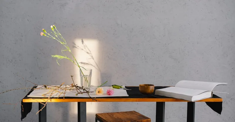 A minimalist scene of a table with flowers, an open book, and wooden bowl, enhanced by natural lighting.