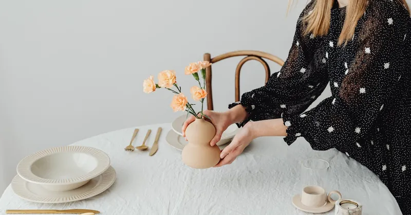 A minimalist table setting featuring a woman arranging flowers in a modern vase.