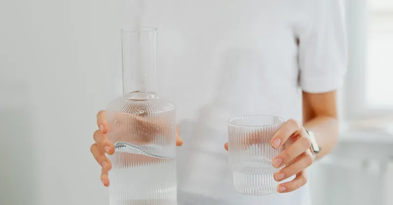 Minimalist image showing hands holding a sleek glass carafe and glass of water.