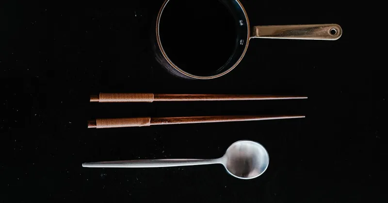 Flat lay of kitchen utensils including chopsticks, pot, and spoon on black surface.