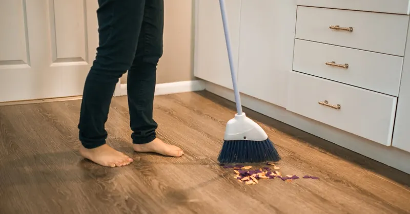 Person sweeping wooden kitchen floor with a broom, showing daily cleaning routine.