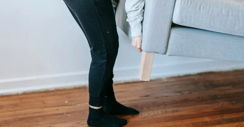 A person lifting a couch in a minimalist living room with wooden floors.