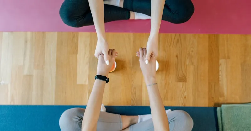 Two people seated on yoga mats holding hands, promoting balance and support.