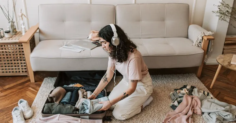 Woman packing suitcase on living room floor, wearing headphones, preparing for a trip.