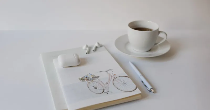 A minimalist setup with a cup of coffee, notepad, pen, and wireless earbuds on a white background.