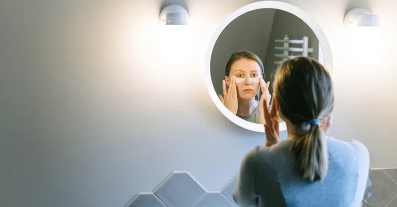 A woman applies under eye patches in a modern bathroom, emphasizing self-care.