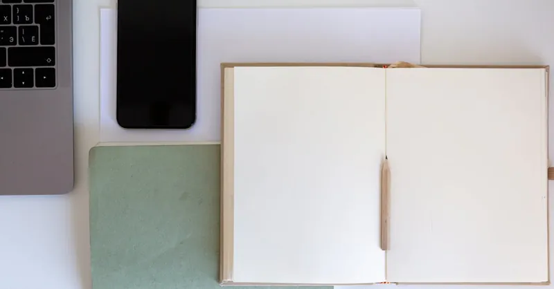 Top view of a minimalist workspace featuring an open notebook, pencil, laptop, and smartphone on a white desk.