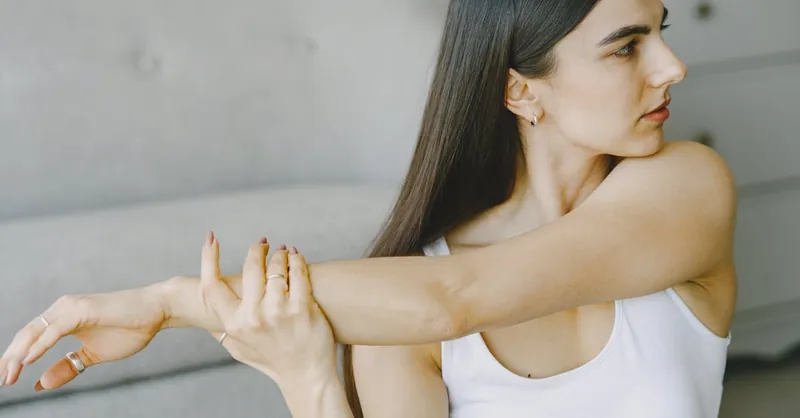 A young woman in a white tank top stretching indoors, focusing on fitness and well-being.