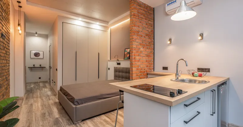 Miniature kitchen with modern appliances and bedroom behind brick partition in small loft style apartment