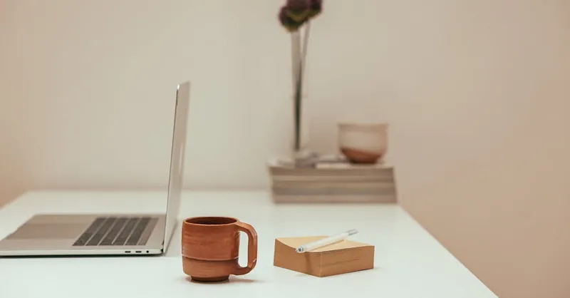A minimalist workspace featuring a laptop, brown mug, pen, and notepad on a white desk.