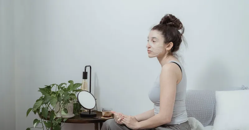 Young woman with a facial mask reflecting on self-care in a minimalist indoor setting.