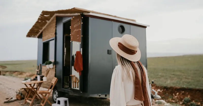 Woman standing by a tiny house on wheels in a rural landscape, capturing the essence of mobile living.